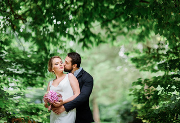 Fototapeta premium Bearded groom kisses bride while they stand under arch of greenery