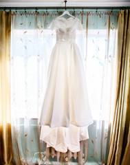 Long wedding dress with lace top hangs before window with beige curtains