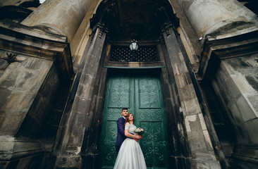 Tall groom hugs bride from behind standing with her before entrance to an old cathedral