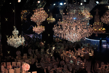 Luxury large crystal chandeliers hang over round dinner tables prepared for the party