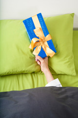 woman hand coming out of blue and green sheets with paper wrapped gift box with ribbon over pillow
