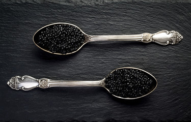 Two vintage silver spoons with black sturgeon caviar on black slate stone background. Top view,...