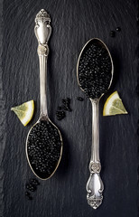 Two vintage silver spoons with black sturgeon caviar on black slate stone background. Top view,...