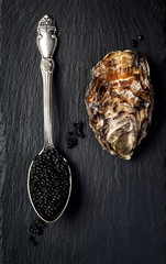 Vintage silver spoon with black sturgeon caviar and oyster on black slate stone background. Top...