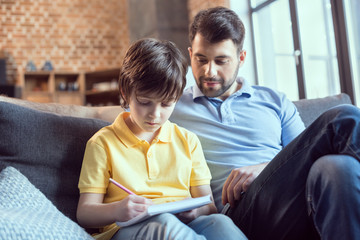Smiling father looking at cute little son doing homework at home