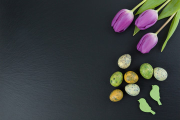 Colorful quail eggs and tulip flowers and green wooden birds on stone table. Top view with copy space