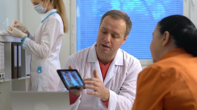 Handsome medical doctor is talking to female and showing her a X-ray picture on tablet while working in his office