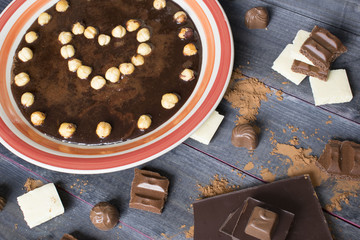 Chocolate composition, a plate with chocolate and hazelnuts in the form of heart,, bar of chocolate, pieces of chocolate and some cocoa on the grey wooden background.