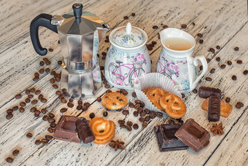 A coffee pot, a sugar bowl, a milk pot, some pieces of chocolate, chocolate sweets, shortbread cookies, coffee beans and star anise on a white wooden background