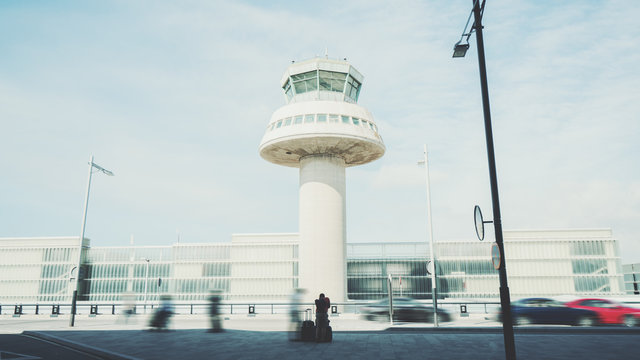 Rear view of tired man with luggage waiting for taxi after his arrival in Barcelona, silhouette of young guy with several suitcases in front of air traffic control tower of modern contemporary airport