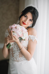 Naklejka premium beautiful and young bride with bouquet of flowers standing near window