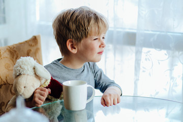Cute little boy holding holding toy lamb and drinking milk or tea from big white cup in sunny morning. © Iryna