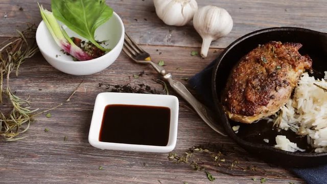 Roasted duck meat on cast iron pan, rice, soy sauce, spices, garlic bulbs on wooden table. Slow pan. Top view.
