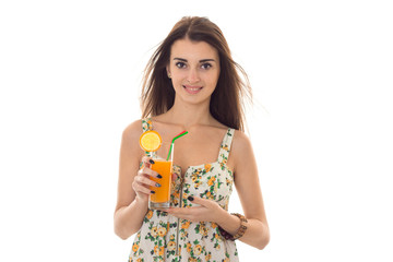 cheerful brunette woman in sarafan with floral patter drinks fresh orange cocktail and smiling on camera isolated on white background