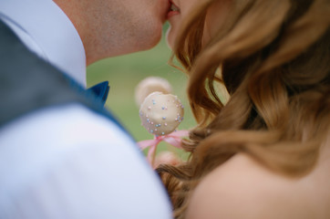 the bride and groom in a blue suit and bow tie are holding cake-pop, kissing in the summer on the nature close up