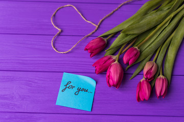 Pink colorful tulips over a purple background, in a flat lay composition with heart and inscription on paper for you.