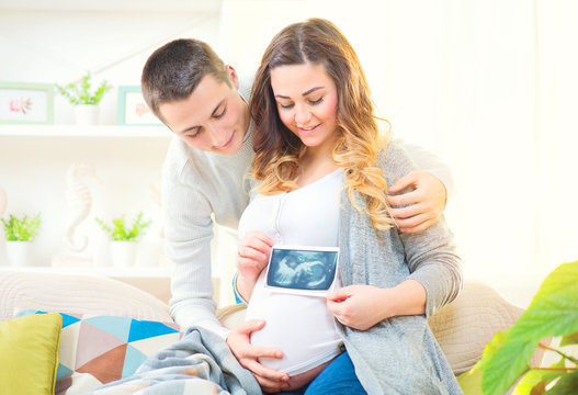 Happy young couple expecting baby. Beautiful pregnant woman and her husband together holding ultrasound picture of their baby