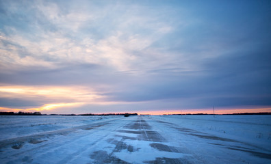 Snow blowing across a gravel back road dividing agriculture snow covered fields in a barren winter landscape at sunset
