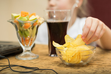 Girl eats potato chips and fruit salad in front of laptop.