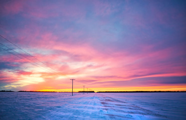 A orange, yellow, and purple sunset over a snow swept countryside gravel road dividing agriculture fields in a barren rural landscape