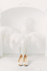 White wedding shoes on the background of the fireplace and ostrich feathers in white interior