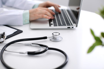Close-up of stethoscope is lying on the table near female doctor typing on laptop computer