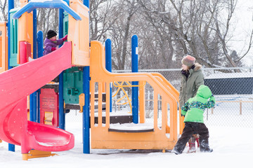 Family at playground in snow, mother watches children playing