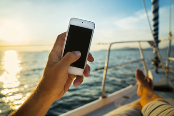 Summer leisure. Rest on a yacht with a phone in his hand. Man lying on the deck and enjoy your smartphone. The guy doing the photo feet on the background seascape and yachts. - Powered by Adobe