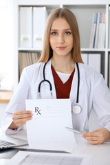 Fototapeta na wymiar Young female doctor fills up medical form and pointing into medical prescription copy space area while looking at the camera. Health care and medicine concept