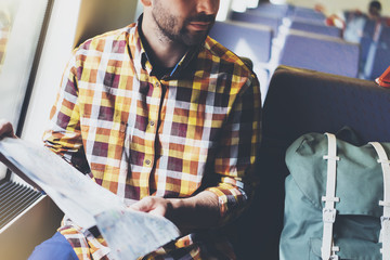 Enjoying travel. Young hipster smile man with backpack traveling by train sitting near the window holding in hand and looking map. Tourist in shirt planing route of railway, railroad transport concept