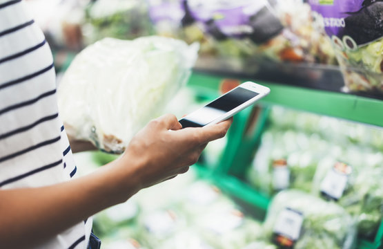 Young woman shopping healthy food in supermarket blur background. Female hands buy products cabagge using smartphone in store. Hipster at grocery using smartphone. Person comparing price of produce