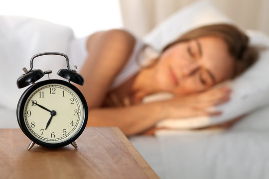 Beautiful young woman sleeping and smiling while lying in bed comfortably and blissfully on the background of alarm clock is going to ring. Sunbeam dawn  on her face.