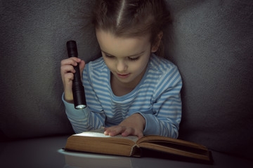 Little girl reading fairy tales book under the covers at the evening with lantern. Cute kid playing before going to sleep, image toned.