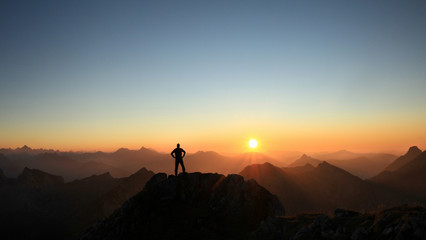 Man reaching summit enjoying freedom and looking towards mountains sunset. - obrazy, fototapety, plakaty