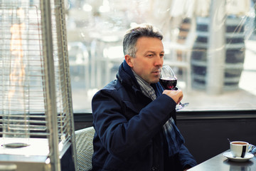 Handsome man in street winter cafe drinking coffe and wine. Male portrait. Attractive confident middle-aged man sitting in city bar, image toned.