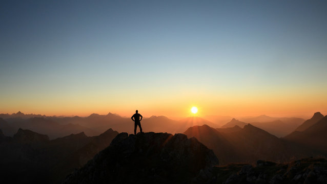 Man reaching summit enjoying freedom and looking towards mountains sunset.