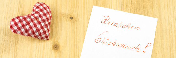 Ein selbstgenähtes Herz und ein weißer Zettel mit der Aufschrift Herzlichen Glückwunsch, Holzhintergrund, Banner,