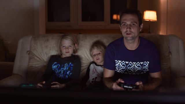 Handsome Caucasian father in blue t-shirt sitting at sofa with his two blonde sons playing console