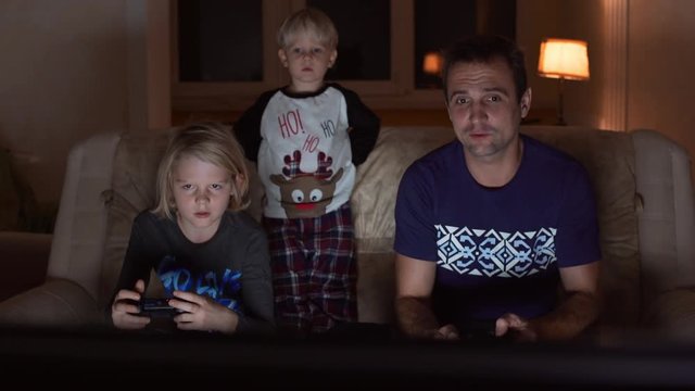 Middle-aged Caucasian father sitting at sofa and playing console with his blonde young son while little one watching