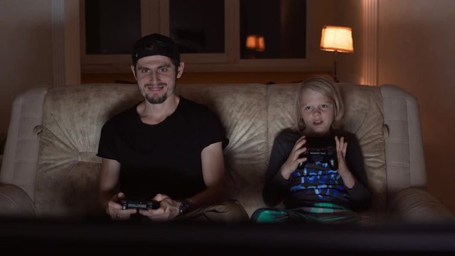 Adult Caucasian male with cap light bristle and black t-shirt sitting at sofa playing console with blonde young boy
