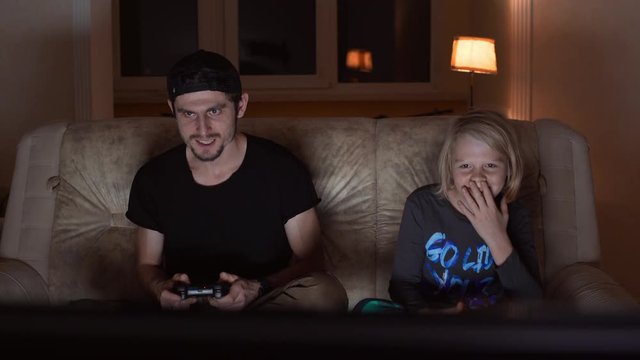 Concentrated adult male in black t-shirt and cap playing console with blonde young boy than leaving