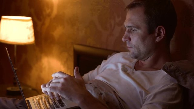 Smiling middle-aged male lying in bed in white t-shirt working on laptop close to torchere in slowmotion