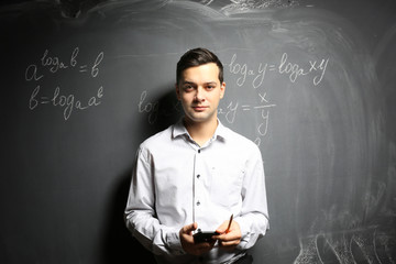 Handsome young teacher with mobile phone standing near blackboard