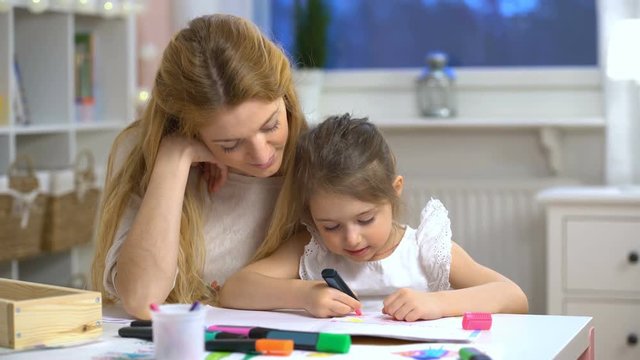 Little girl draws in the nursery. Mother looking how her child daughter drawing a picture
