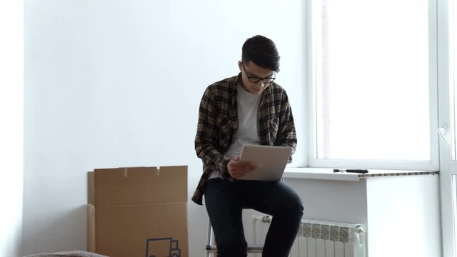 Smiling young man looking for some decoration ideas on a tablet. Moving into a new apartment. Cardboard boxes in the background.