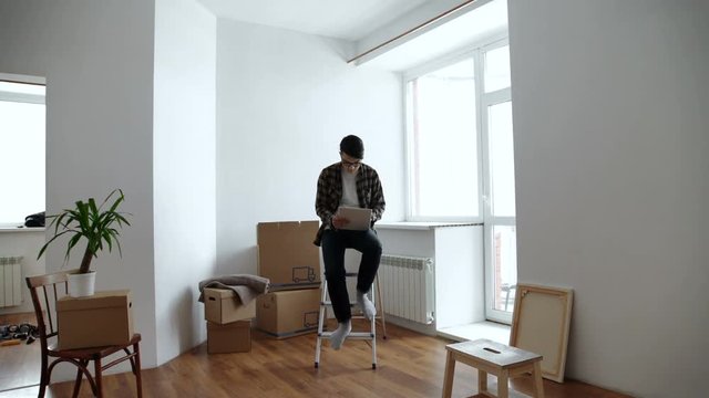 Smiling young man looking for some decoration ideas on a tablet. Moving into a new apartment. Cardboard boxes in the background.