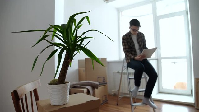 Smiling young man looking for some decoration ideas on a tablet. Moving into a new apartment. Cardboard boxes in the background.