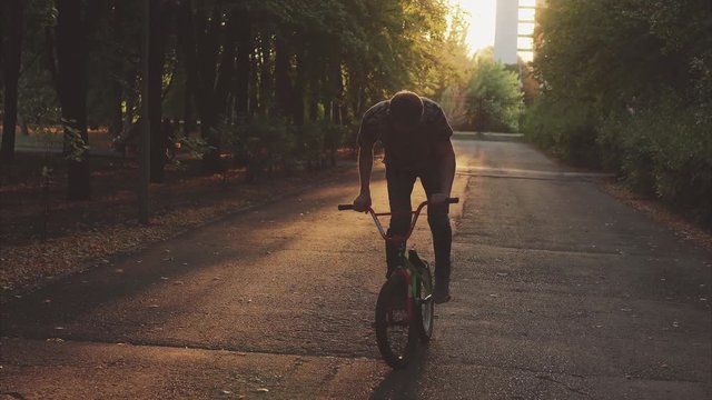 Guy dressed in a free style riding in the park with tall trees on a bicycle BMX. He is confident on it, hold the handles on the steering wheel.