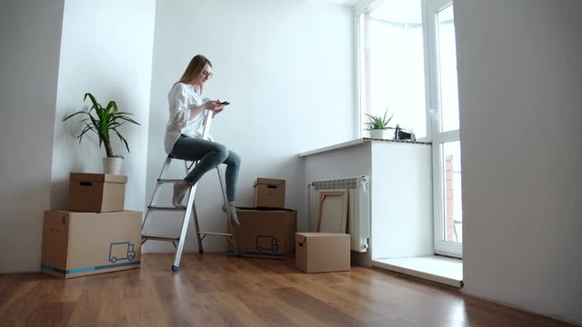 Attractive young woman is moving, standing among cardboard boxes, using a smartphone and smiling