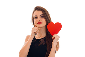 young charming brunette woman in stylish black dress with red heart in her hands looking away isolated on white background